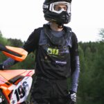 Motocross-Fahrer in Schutzkleidung steht neben einem orangefarbenen Motorrad und trägt ein Trikot mit Sponsorenlogos von Elektro Reichl.