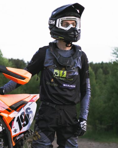 Motocross-Fahrer in Schutzkleidung steht neben einem orangefarbenen Motorrad und trägt ein Trikot mit Sponsorenlogos von Elektro Reichl.