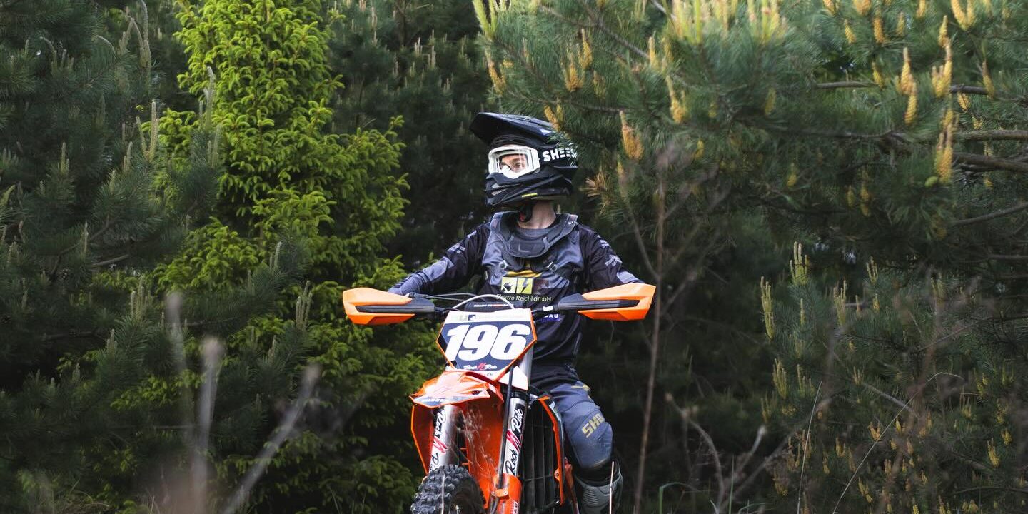 Motocross-Fahrer sitzt auf einem Motorrad in einer bewaldeten Umgebung und trägt Bekleidung mit Sponsorenlogos von Elektro Reichl.