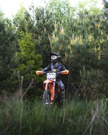 Motocross-Fahrer sitzt auf einem Motorrad in einer bewaldeten Umgebung und trägt Bekleidung mit Sponsorenlogos von Elektro Reichl.