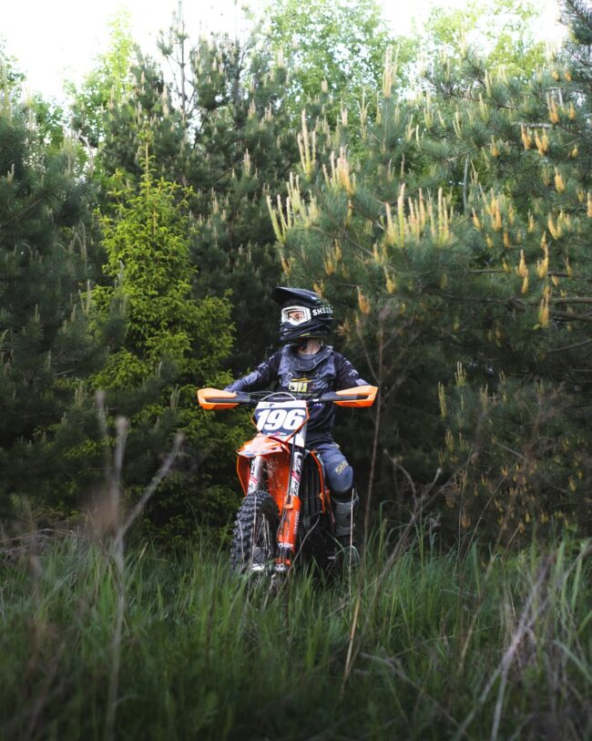 Motocross-Sport mit regionalem Sponsoring Motocross-Fahrer sitzt auf einem Motorrad in einer bewaldeten Umgebung und trägt Bekleidung mit Sponsorenlogos von Elektro Reichl.