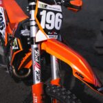 Frontansicht eines orangefarbenen Motocross-Motorrads mit Startnummer und Sponsorenlogo von Elektro Reichl auf der Starttafel.