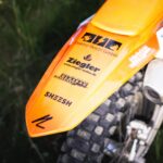 Detailaufnahme des hinteren Kotflügels eines orangefarbenen Motocross-Motorrads mit mehreren Sponsorenaufklebern, darunter Elektro Reichl.