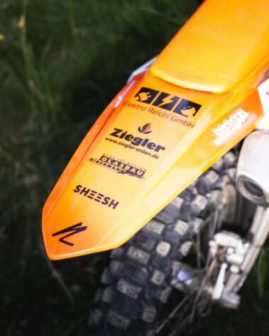 Detailaufnahme des hinteren Kotflügels eines orangefarbenen Motocross-Motorrads mit mehreren Sponsorenaufklebern, darunter Elektro Reichl.