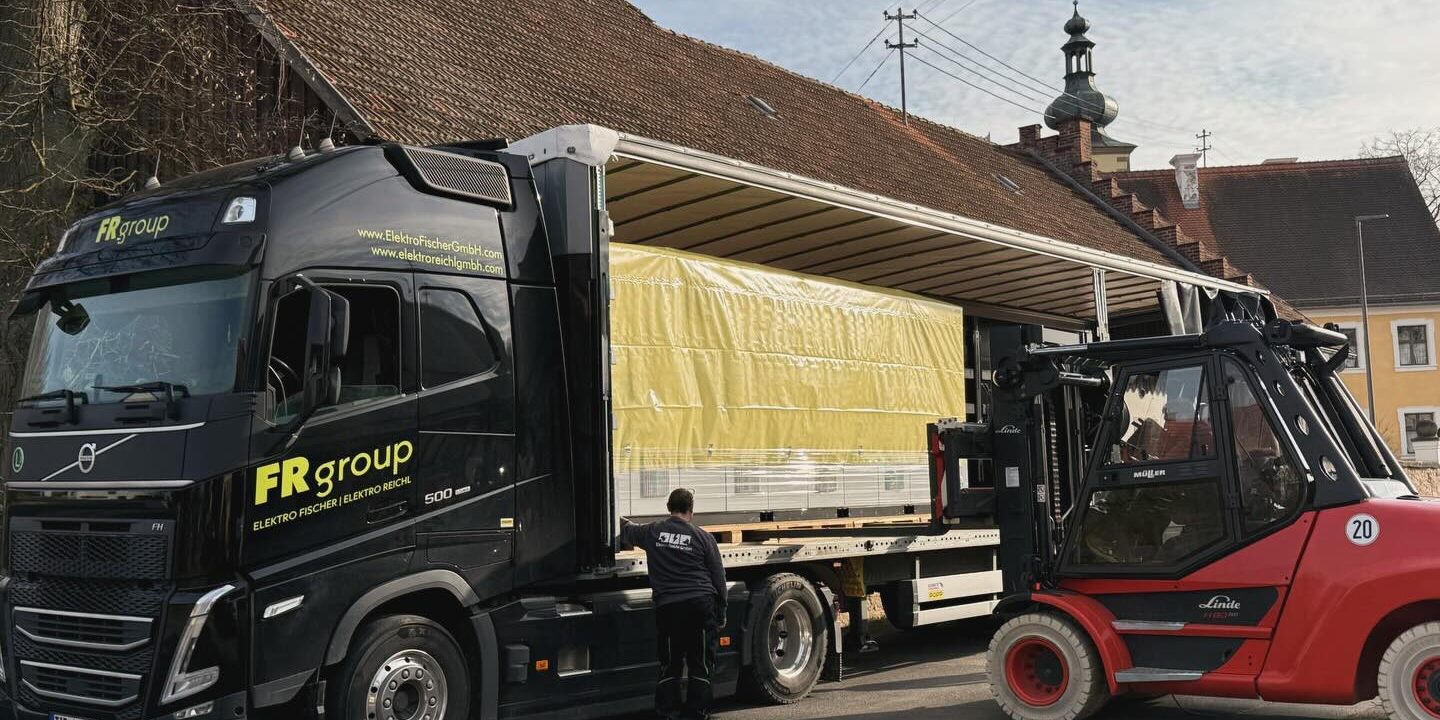 Mitarbeiter und Gabelstapler verladen einen großformatigen Schaltschrank von einem Transportfahrzeug.
