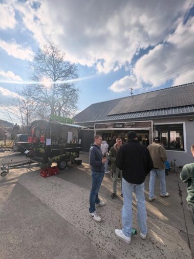 Besucher treffen sich vor einer mobilen Getränkebar und einem Verkaufsstand auf dem Firmengelände bei sonnigem Wetter.