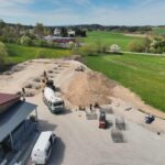 Drohnenaufnahme einer Baustelle mit Aushubarbeiten, Fundamentgruben und Baufahrzeugen zur Vorbereitung eines Neubaus.