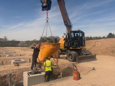 Arbeiter und Bagger unterstützen die Betonage eines Fundaments. Die Aufnahme zeigt einen wichtigen Schritt der Bauausführung.